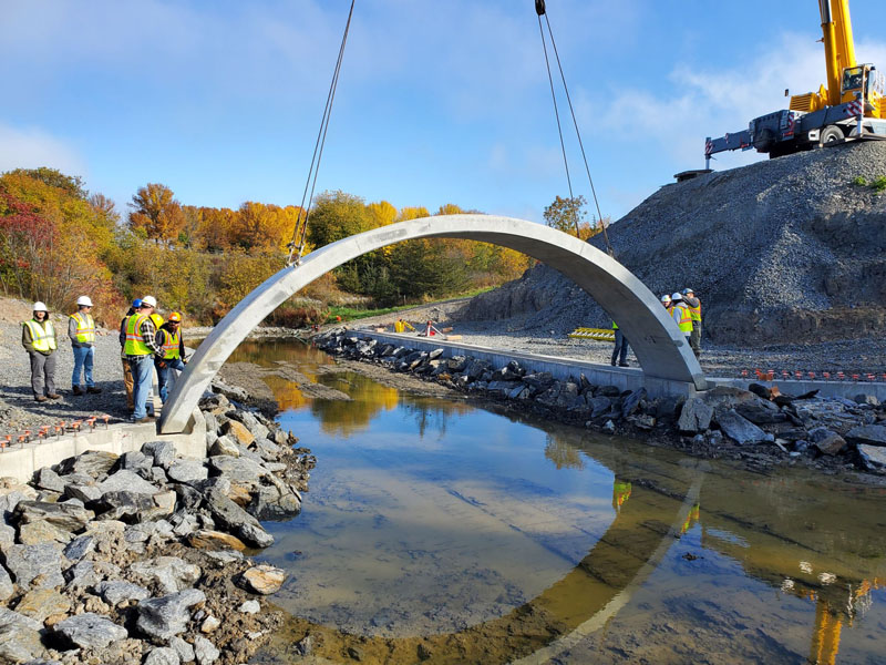 crane carrying arched piece of concrete surrounded by construction workers